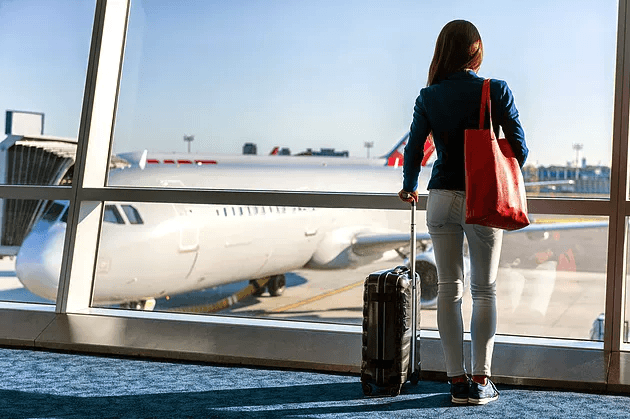 Traveler standing at an airport window with luggage, looking toward an airplane.