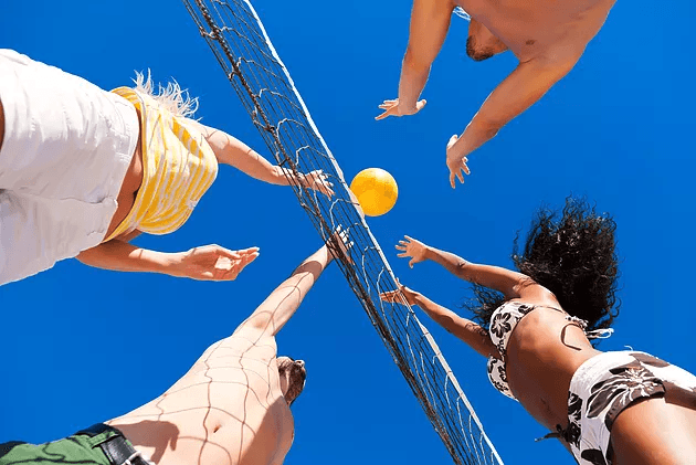People playing beach volleyball, reaching to hit the ball against a bright blue sky.