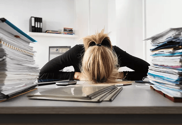 Person resting face-down on a desk surrounded by stacks of papers.