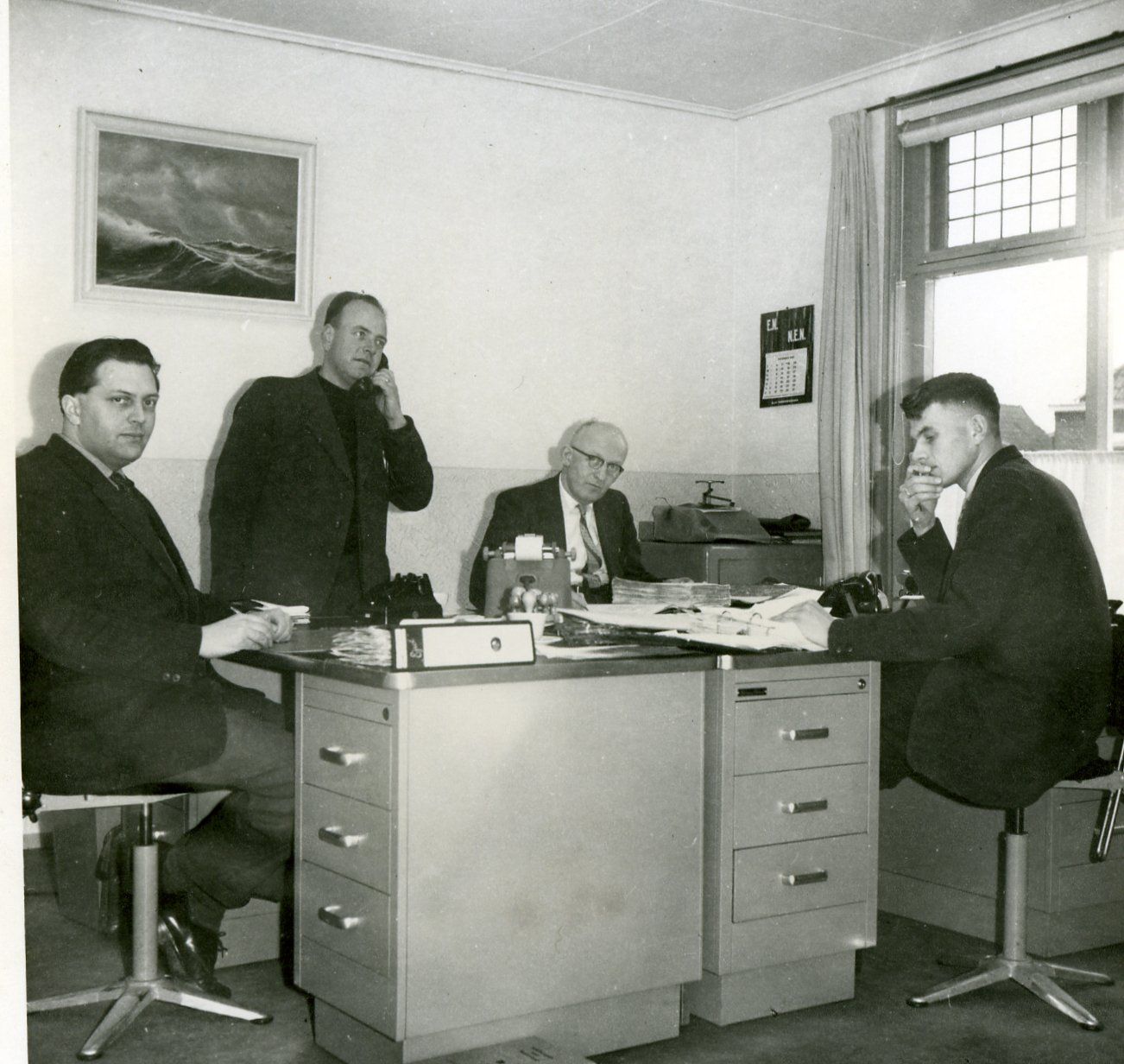 Vier Männer in einem Büro, zwei telefonieren, einer sitzt am Schreibtisch, der vierte steht. Schwarz-Weiß-Foto.