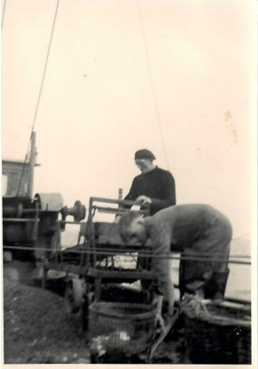 Zwei Männer auf einem Boot, einer an einer Maschine, der andere beim Sortieren eines Netzes, beide in Arbeitskleidung.