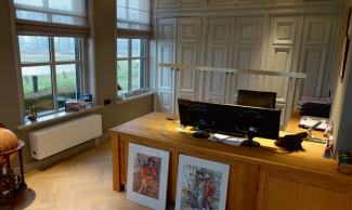 Office with large wooden desk, dual monitors, framed art, and large windows.