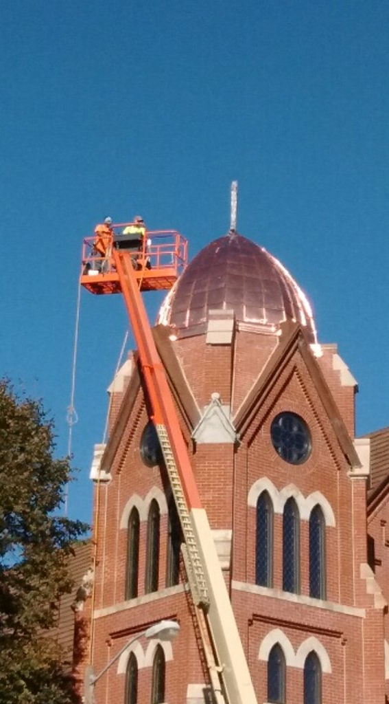 Workers on a lift, repairing copper dome on brick building with a cross under a blue sky.