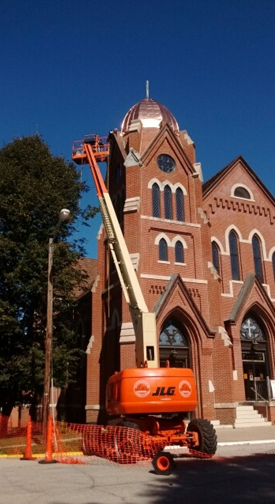 An orange JLG lift next to a red brick church with copper dome. Blue sky.