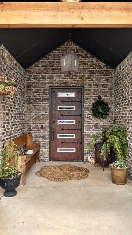 Rustic brick entryway with a brown door, bench, plants, and a woven doormat.
