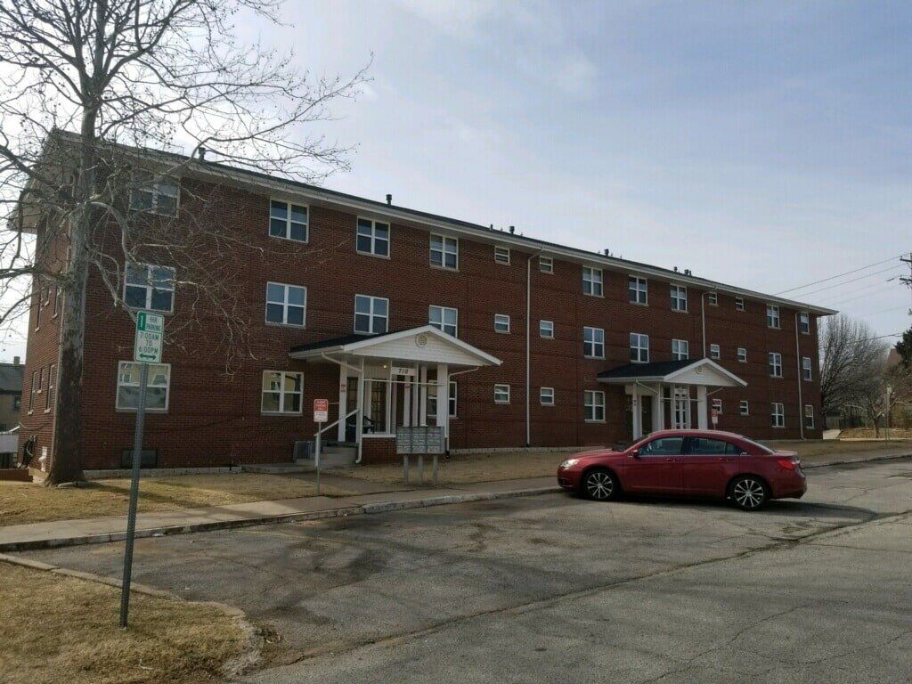 Two-story brick apartment building with multiple windows. A red car is parked in front.