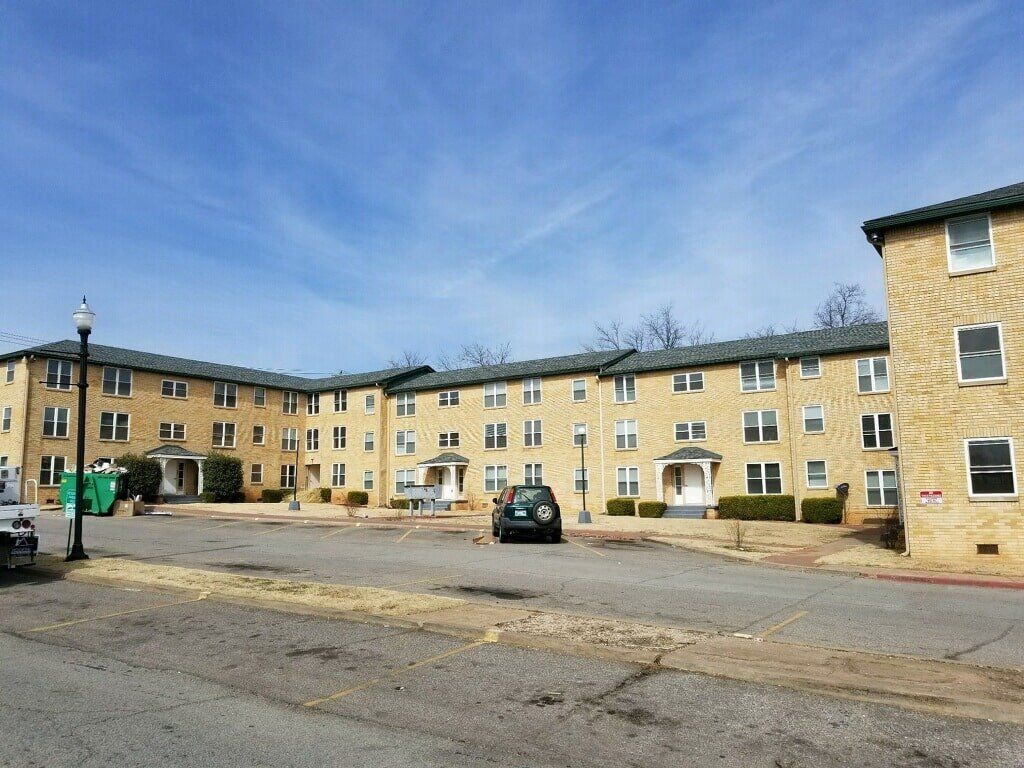 Multi-story beige apartment building with dark roof under a blue sky, car parked in front.