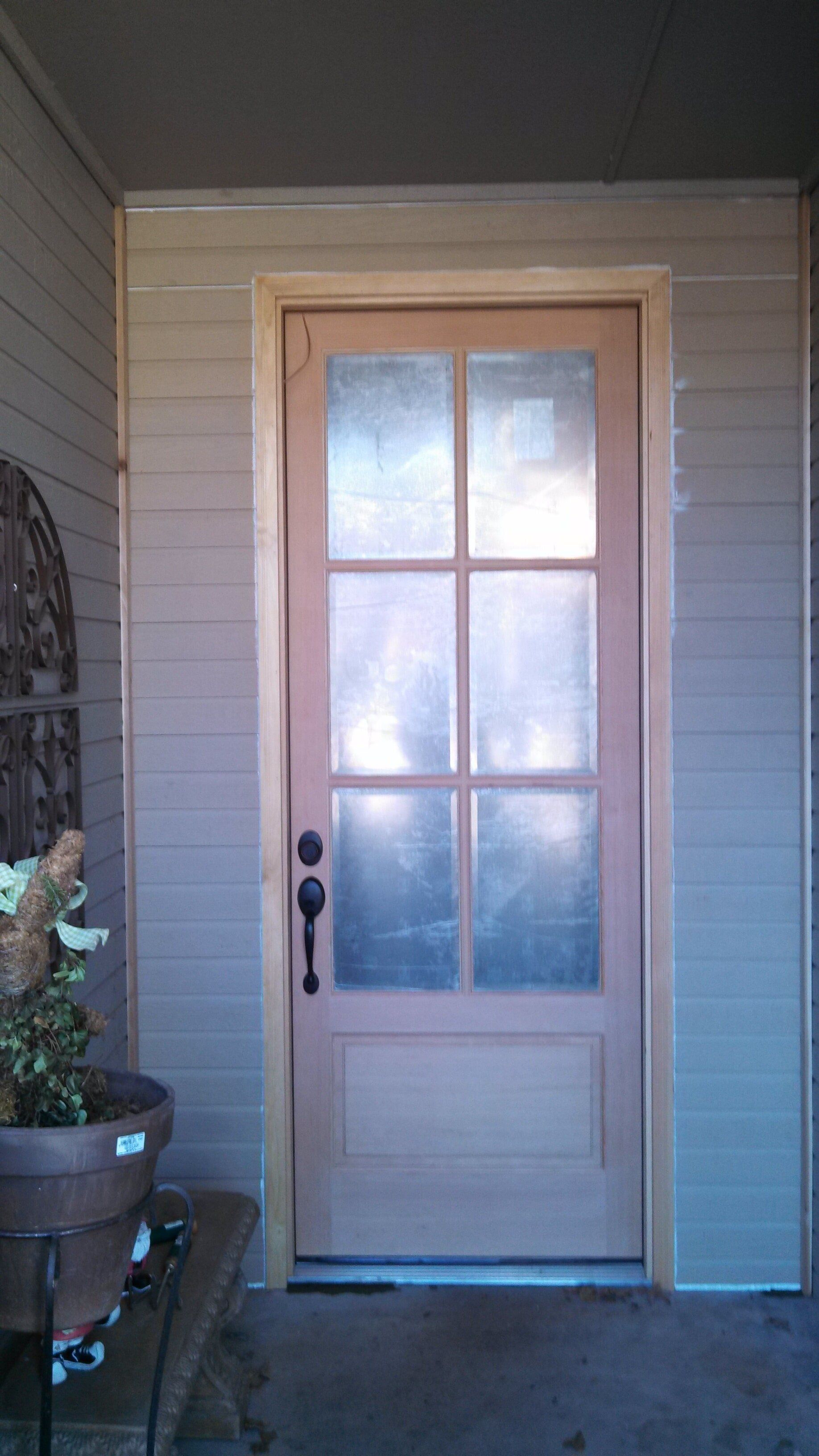 Wooden door with glass panes, framed by light siding and trim. A potted plant sits on the left.