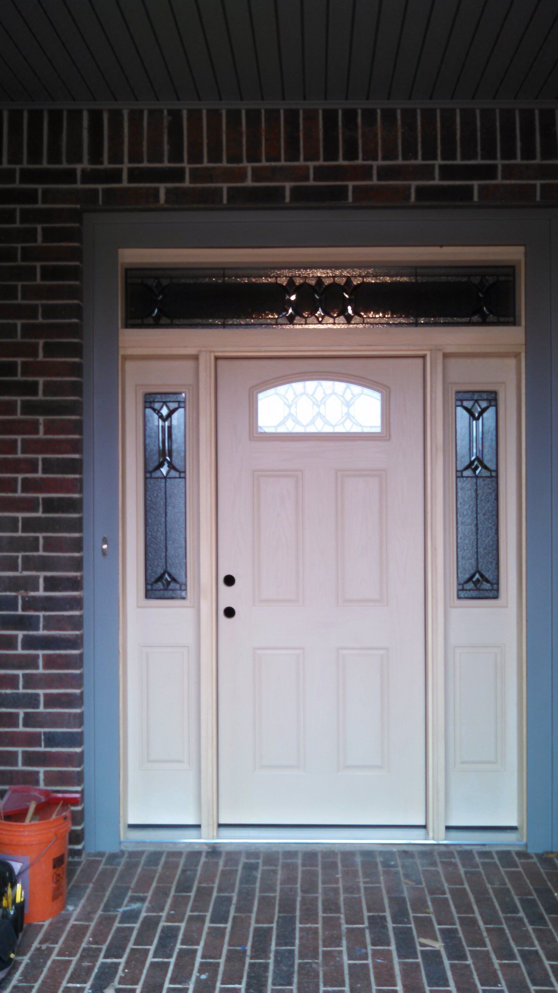 Beige front door with sidelights and transom, set in a brick and gray-painted frame.