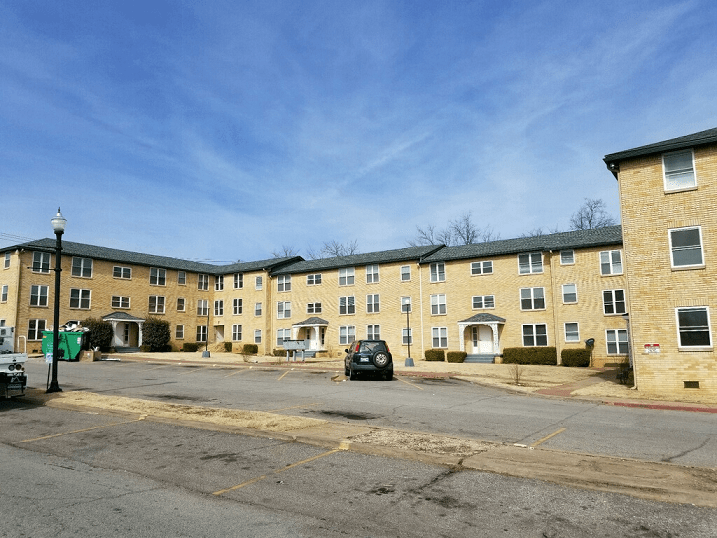 Yellow brick apartment buildings under a blue sky, with a car parked in front and a trash bin on the left.