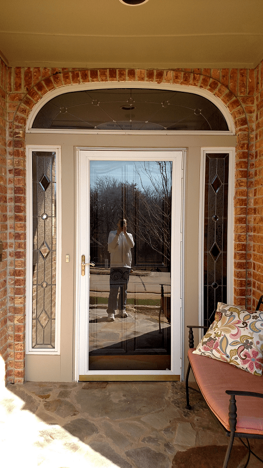 Front door with sidelights and arched transom, reflecting person taking photo; brick facade.