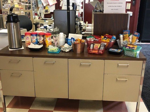 Coffee station: dispenser, snacks, cups, and creamers on a beige cabinet with drawers, red and white checkered floor.