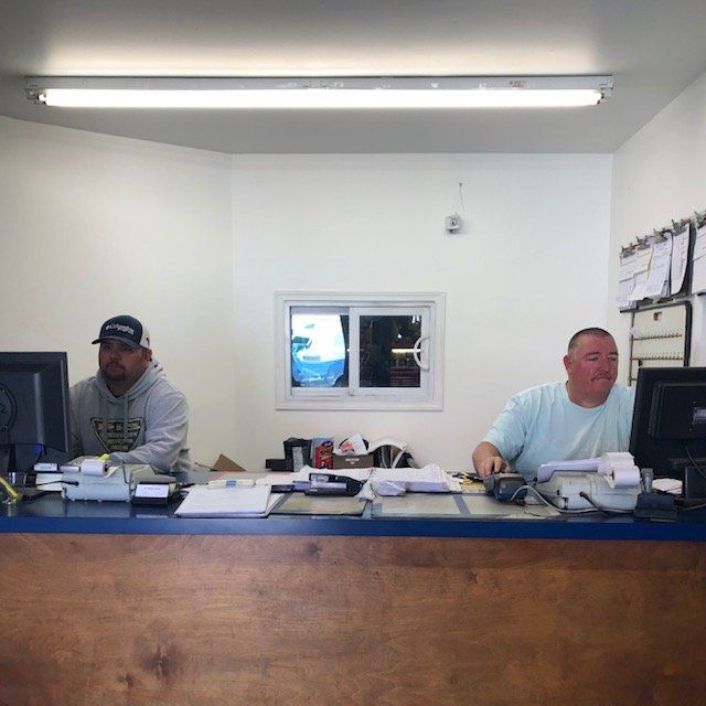 Two men work at a counter in an office. One in a gray hoodie, the other in a teal shirt. White walls, a small window.