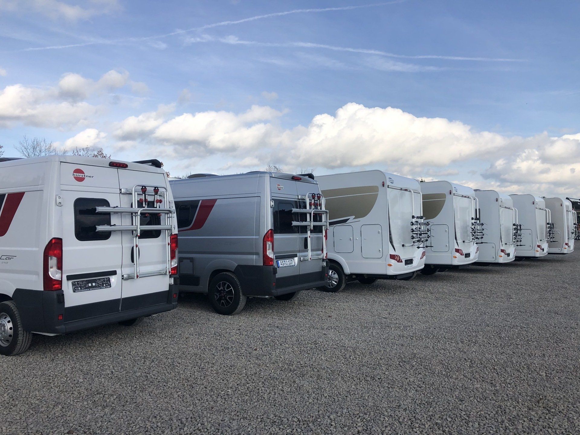 A row of white vans are parked in a gravel lot.