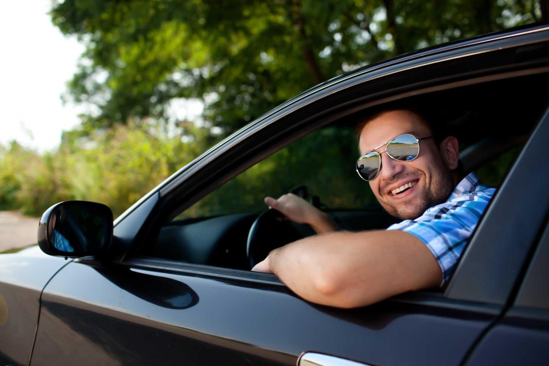 A man wearing sunglasses is driving a car and smiling.