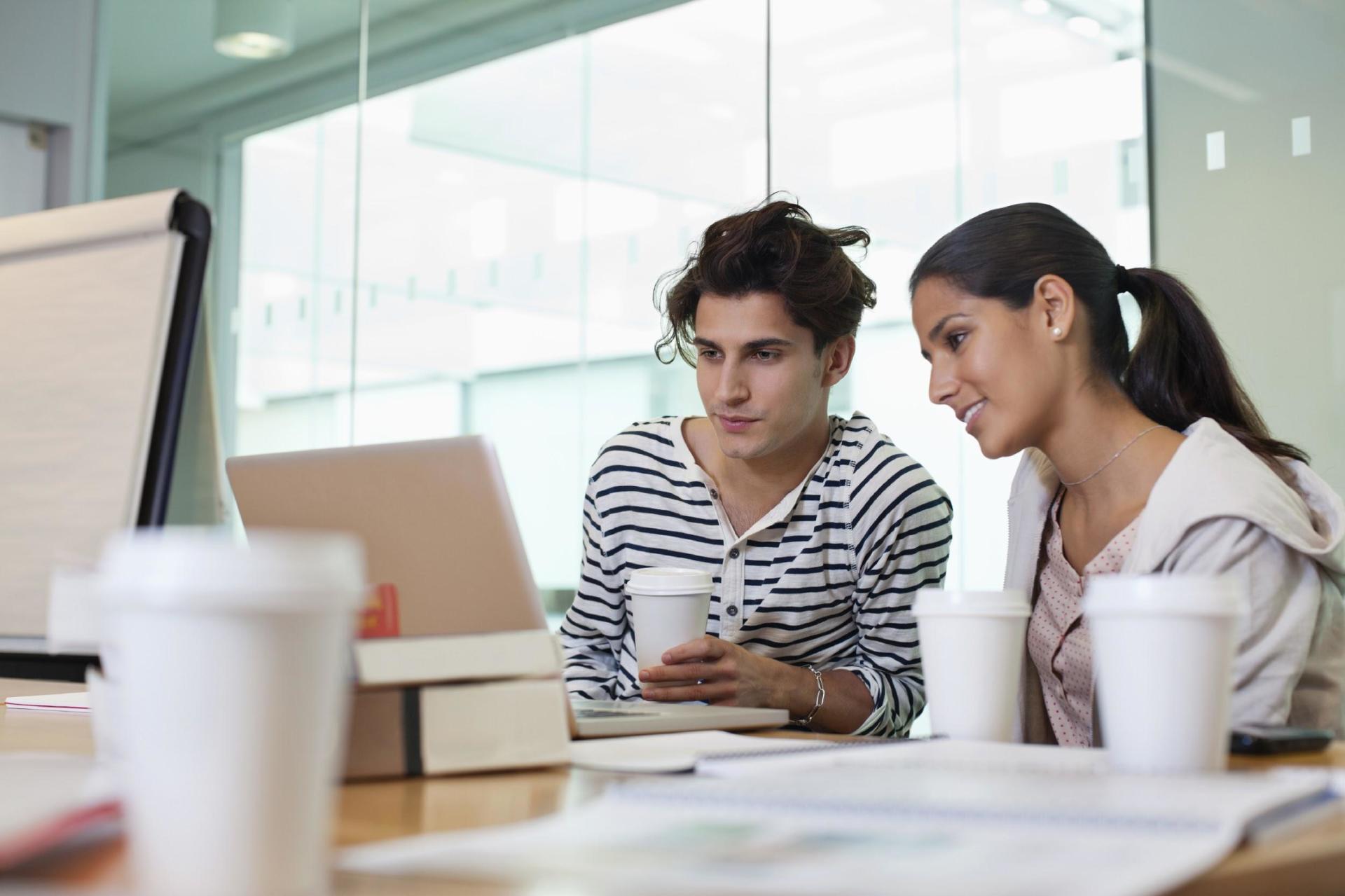 A man and a woman are sitting at a table looking at a laptop.