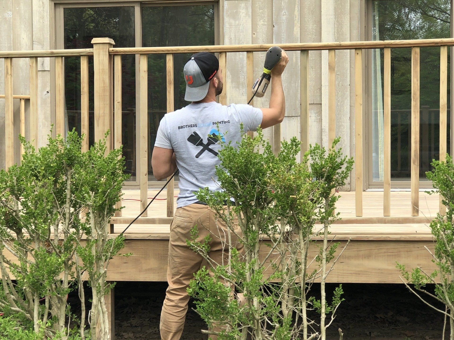 A person in a gray t-shirt uses a power sander to smooth the wooden railing of an outdoor deck.