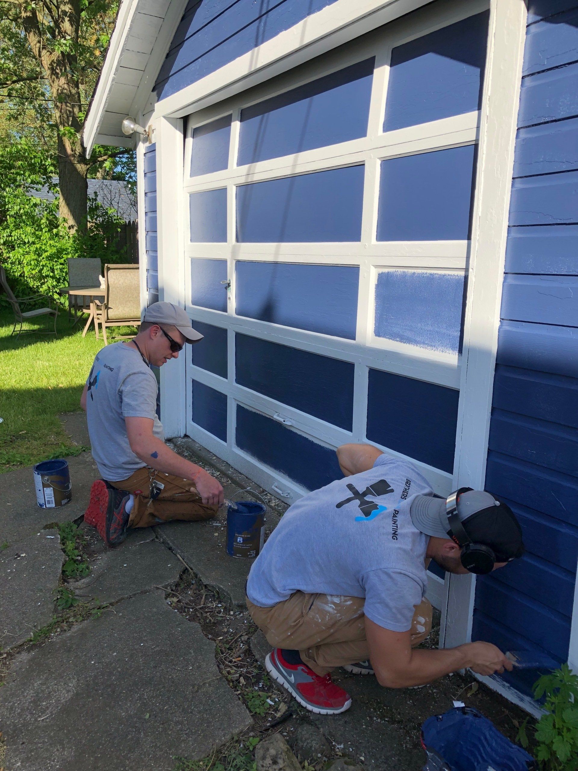 Two people in gray shirts kneeling and painting the base of a blue garage door white.