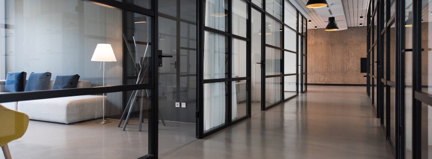 Modern office hallway featuring glass-walled partitions, dark metal frames, concrete floors, and a bright corner lounge.