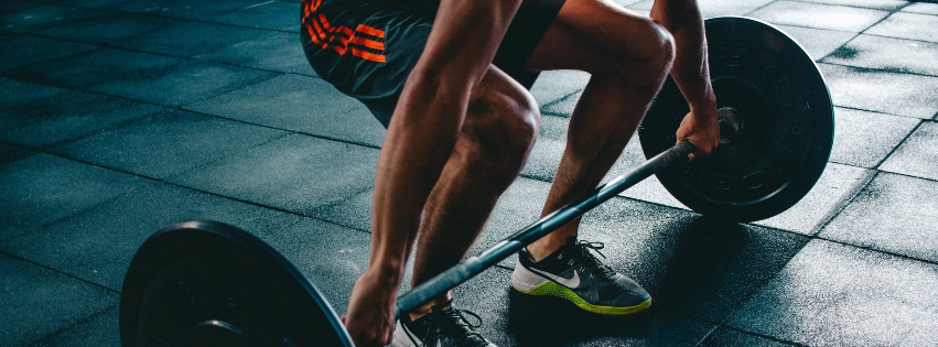 A person in shorts and athletic shoes gripping a barbell on a gym floor, preparing to perform a deadlift.