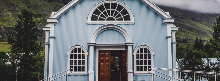 A pale blue wooden church with white trim, arched windows, and a semi-circular fanlight above an open front door.