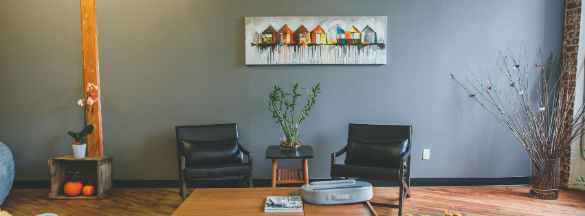Two black leather armchairs sit against a gray wall with a horizontal painting above and a small table between them.