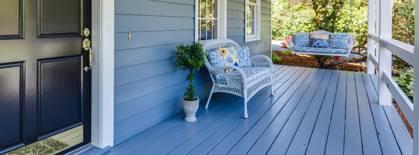A blue porch with a dark front door, a white wicker chair with patterned cushions, and a hanging porch swing.