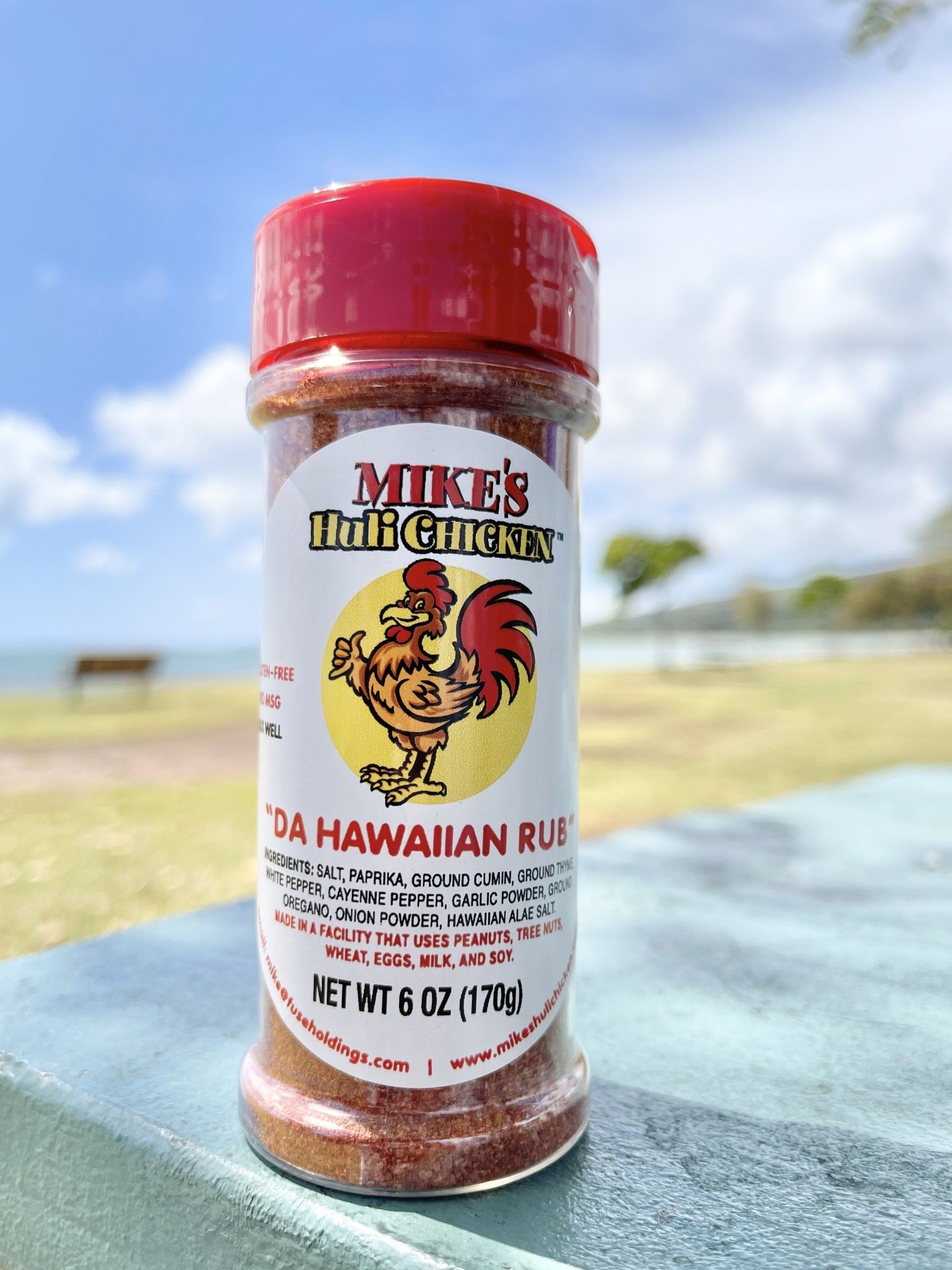 A spice jar of Mike's Ono Chicken Hawaiian Rub sits on a ledge outdoors, with a beach and blue sky in the background.