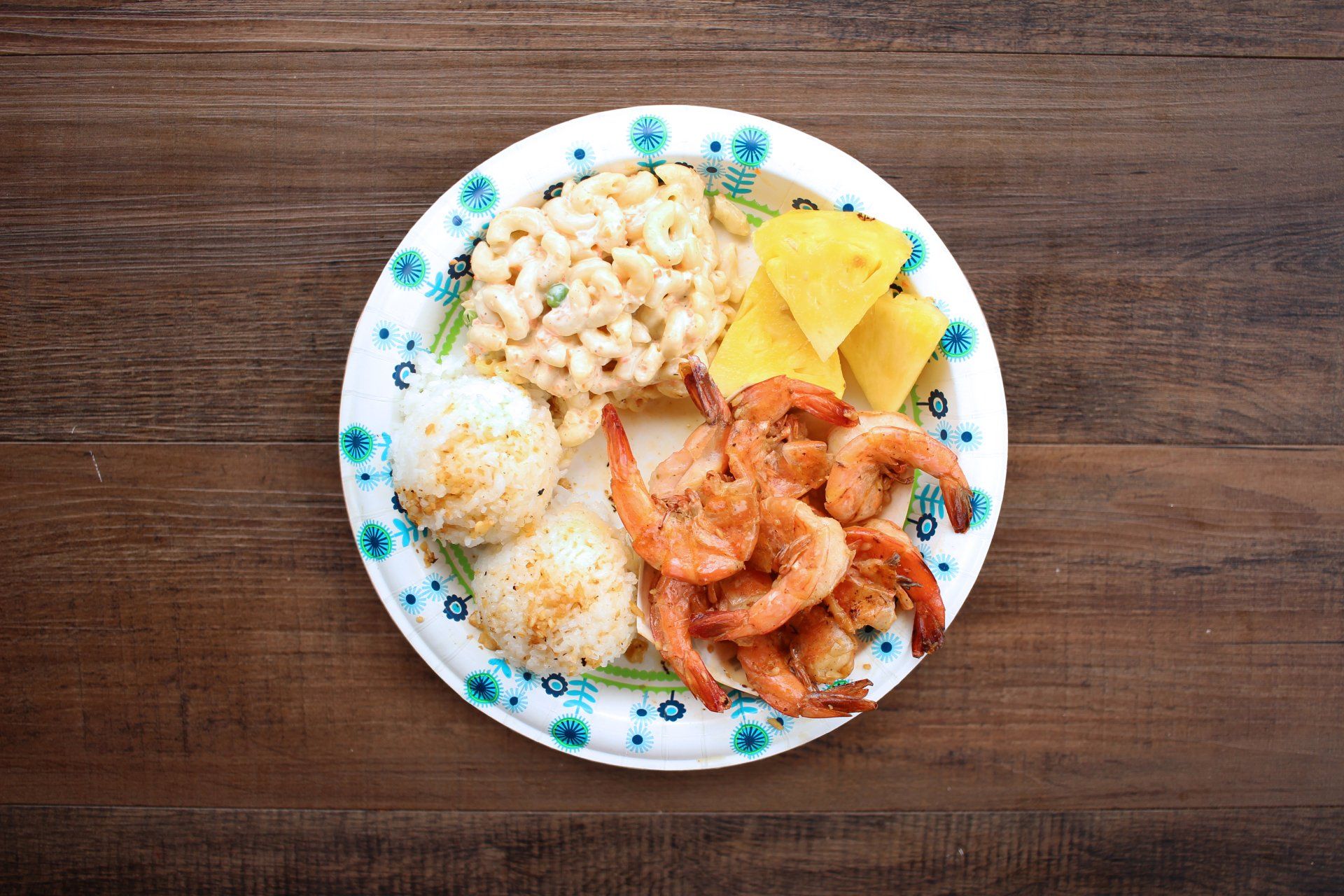 Plate with shrimp, macaroni salad, pineapple, and rice on a wooden surface.