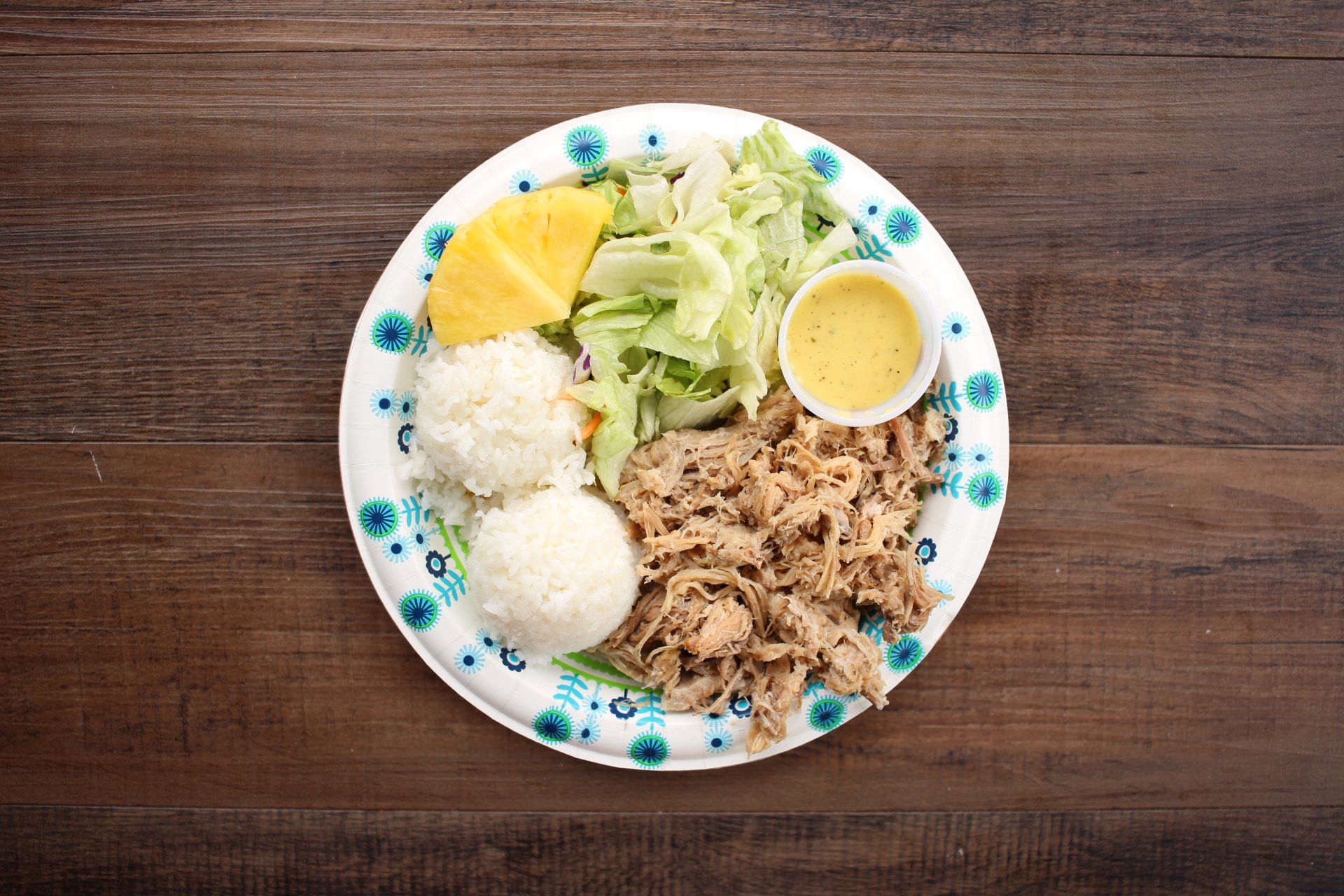 Plate of Hawaiian food with pulled pork, rice, salad, pineapple, and dressing on a wooden table.