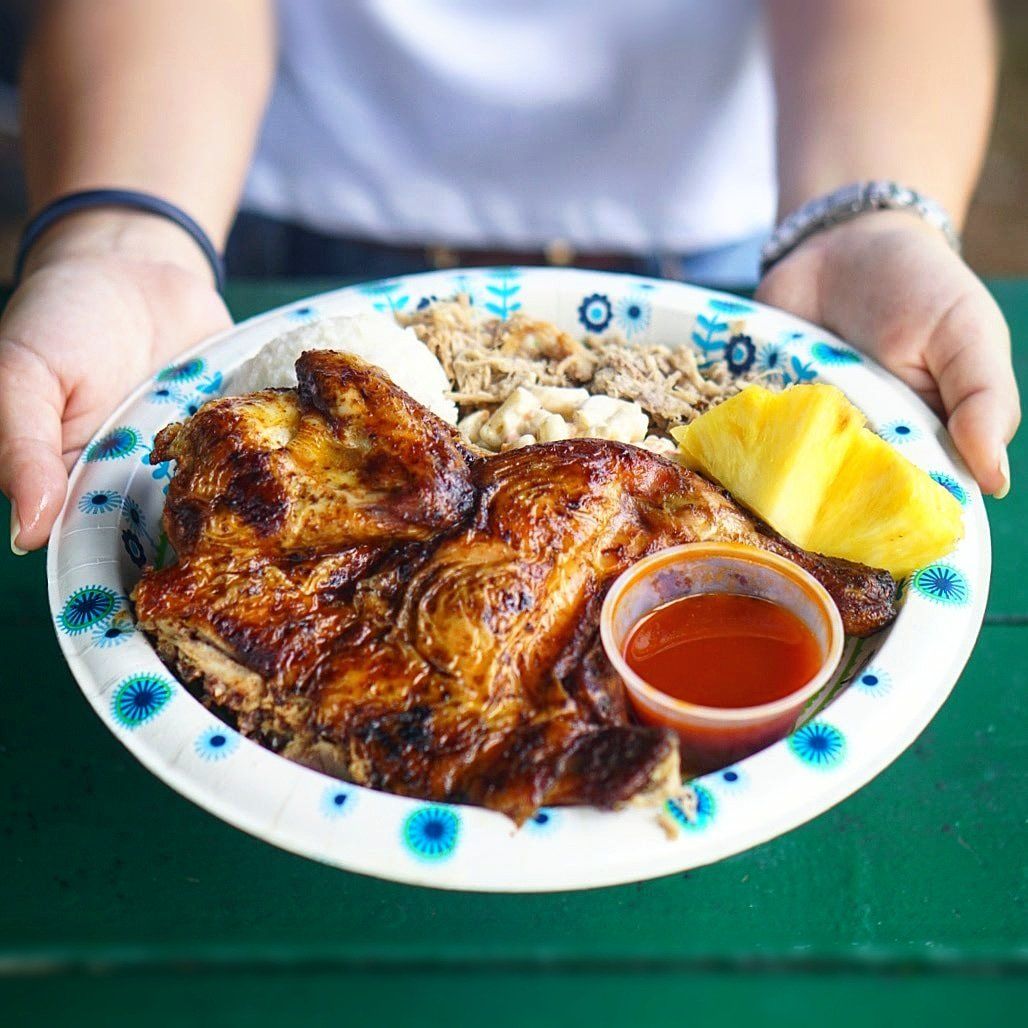 Plate of grilled chicken, mac salad, and pineapple held by a person. Red sauce in a small container is visible.
