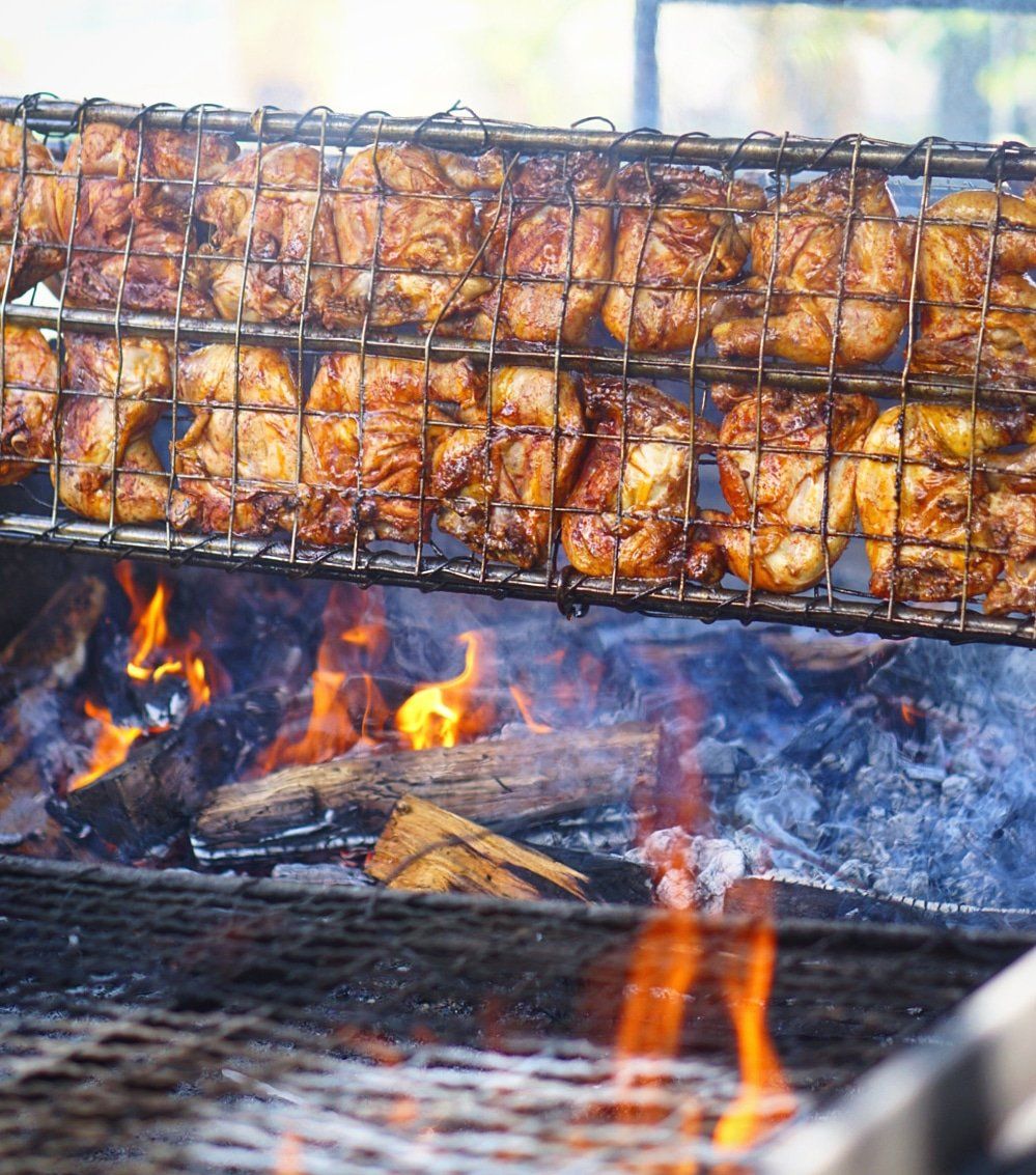 Grilled chicken pieces on a metal rack over a wood fire. The chicken has a crispy, browned appearance.