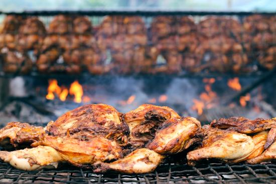 Grilled chicken, close-up of several pieces on a grill with many chickens cooking in the background. Flames and smoke visible.