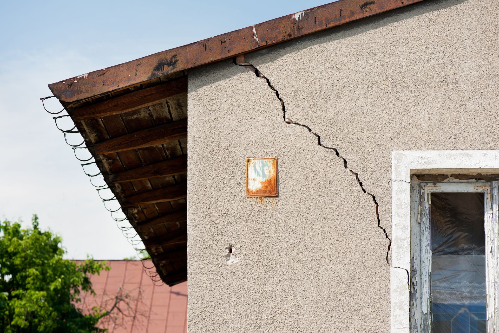 Risse in der Stuckfassade eines Gebäudes mit verrostetem Dach und einem vernagelten Fenster.