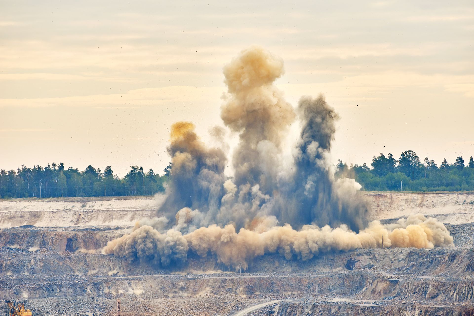 Explosion in einem Steinbruch, eine Staub- und Schuttwolke wird aufgewirbelt, im Hintergrund eine Baumreihe.