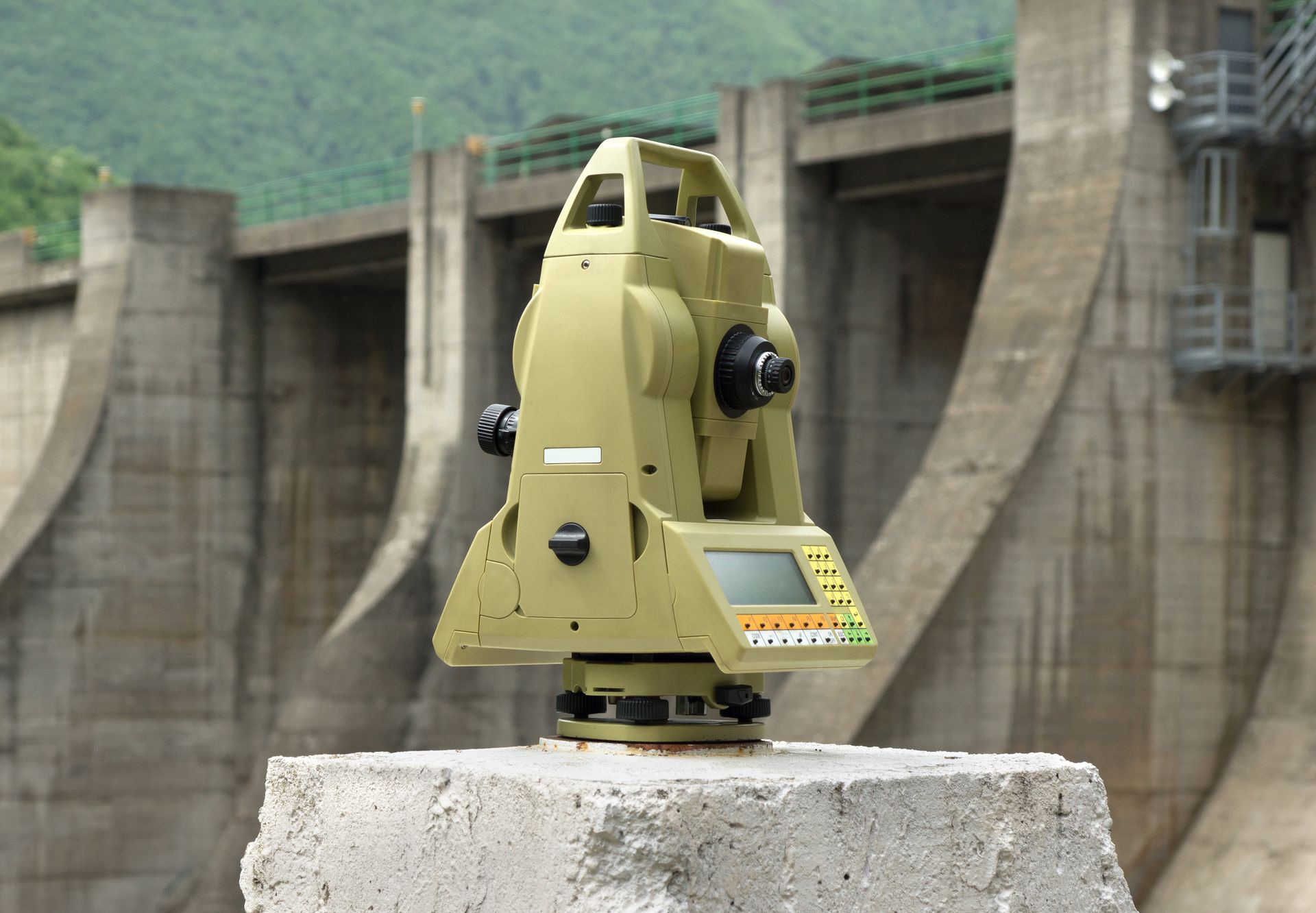Vermessungsinstrument auf einem Betonsockel, im Hintergrund ein Staudamm und Berge.