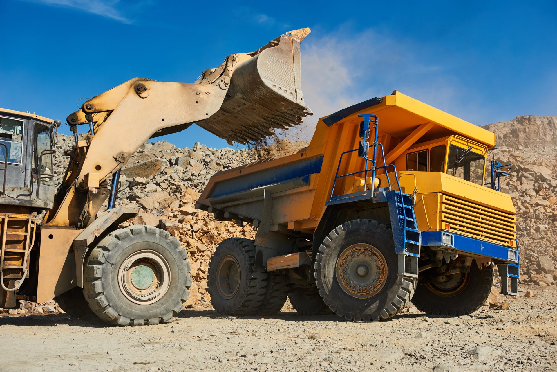 Ein gelber Radlader schaufelt unter blauem Himmel Erde in einen gelben Muldenkipper auf einer Baustelle.