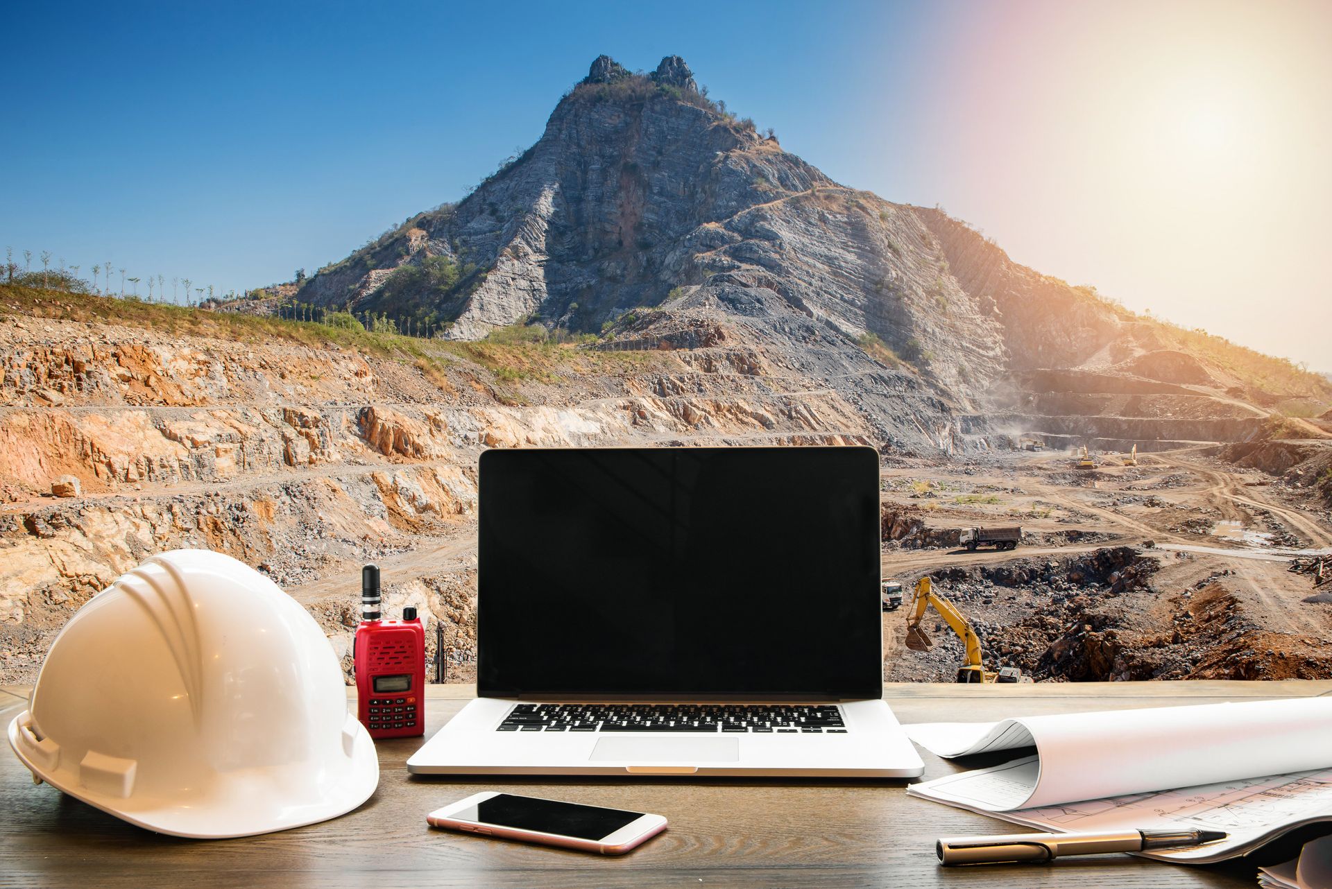 Weißer Schutzhelm, Laptop, Walkie-Talkie und Baupläne auf einem Schreibtisch; im Hintergrund ein Steinbruch in den Bergen.