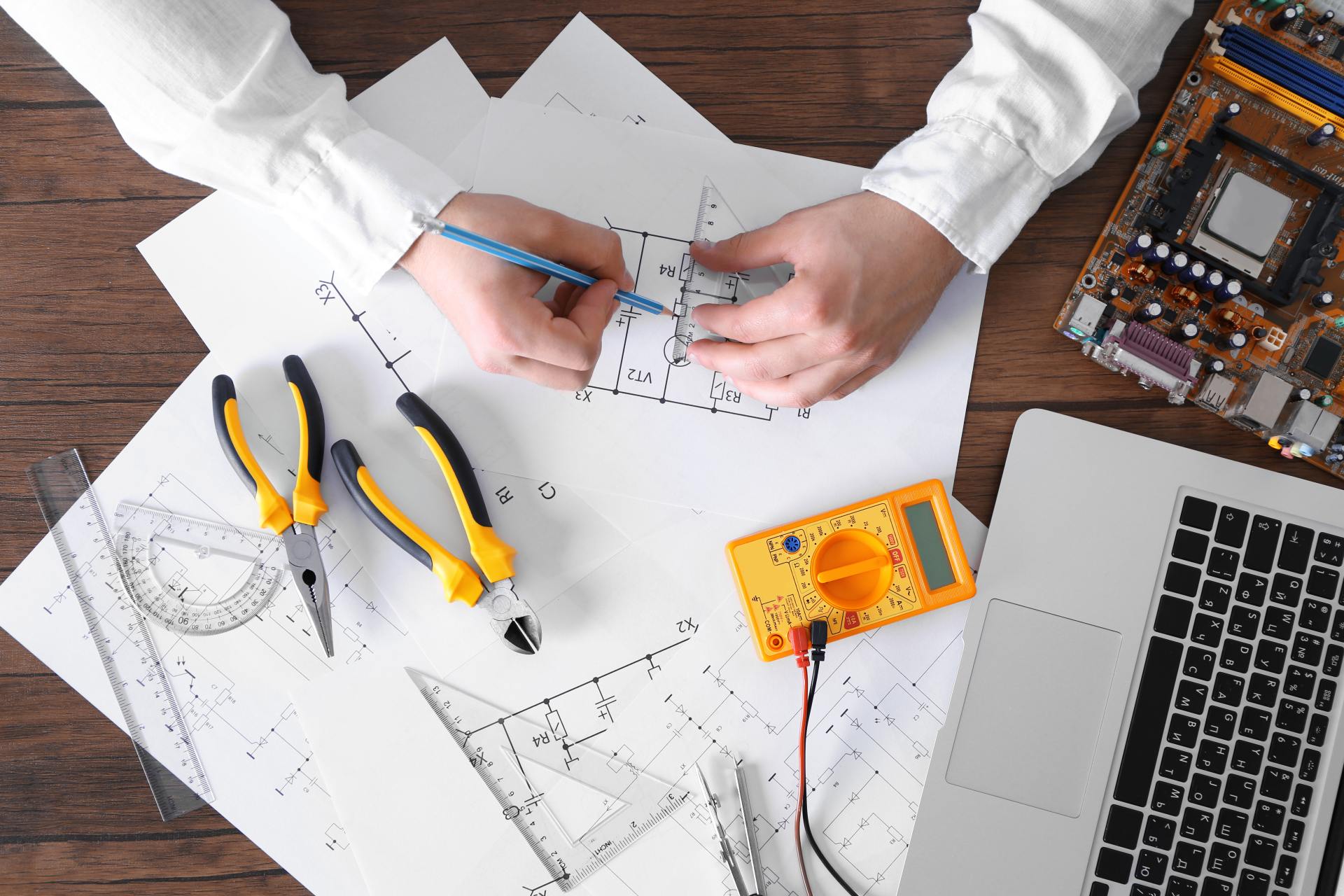 Mani che disegnano uno schema elettrico su carta con strumenti, multimetro, computer portatile e componenti elettronici su una scrivania