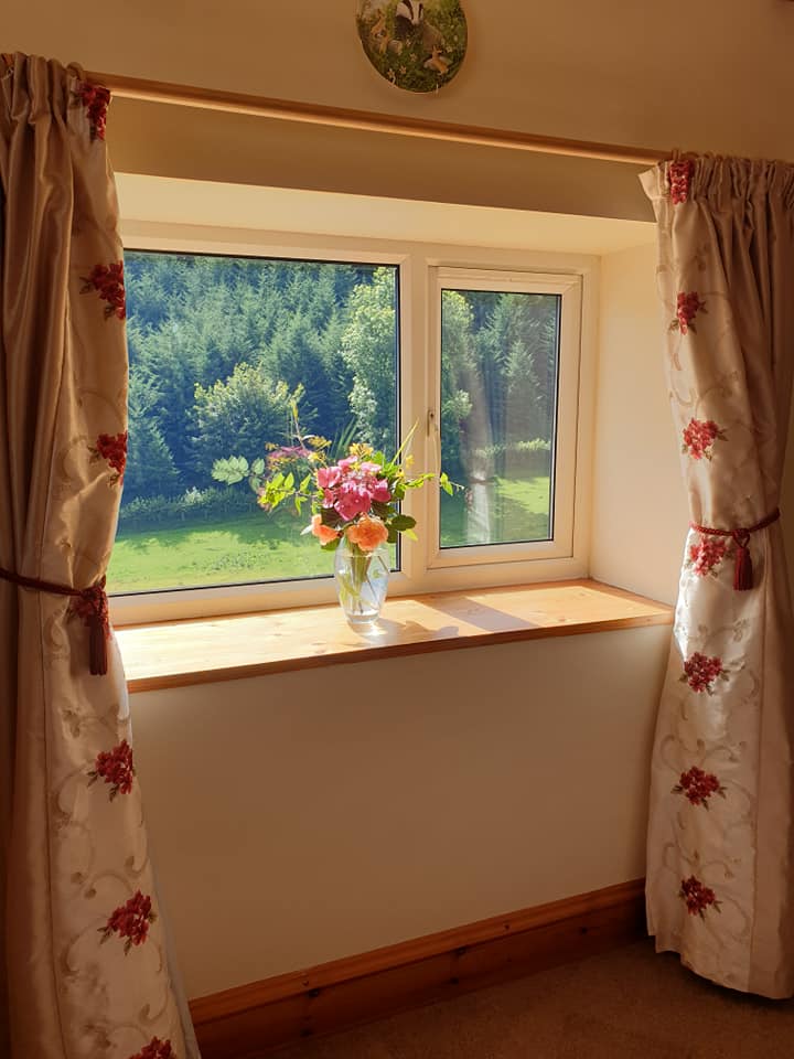 Window with flower arrangement, overlooking a green landscape. Beige curtains with floral pattern.