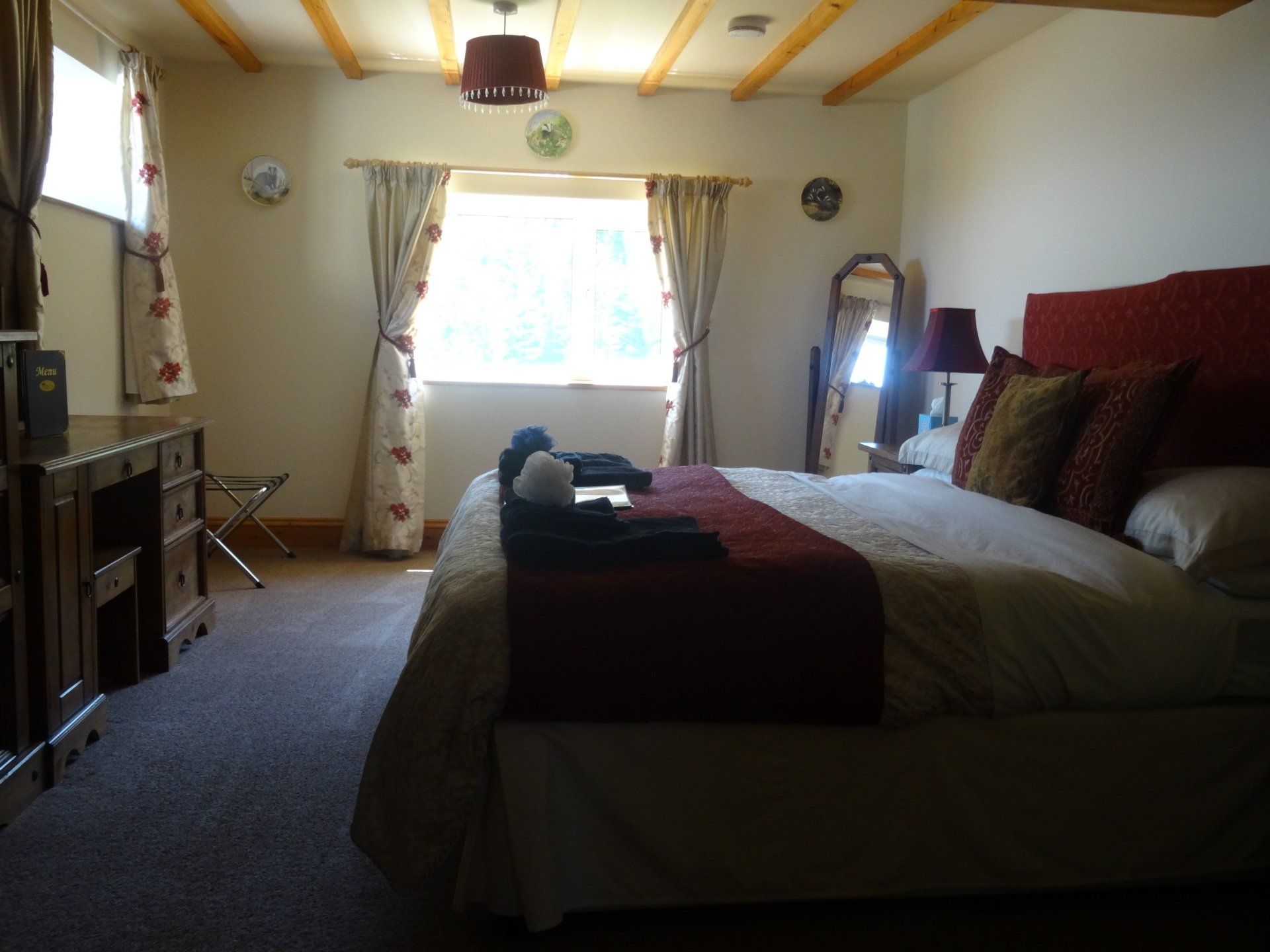 Bedroom with a bed, dresser, window, and wood-beam ceiling. Neutral colors with red accents.