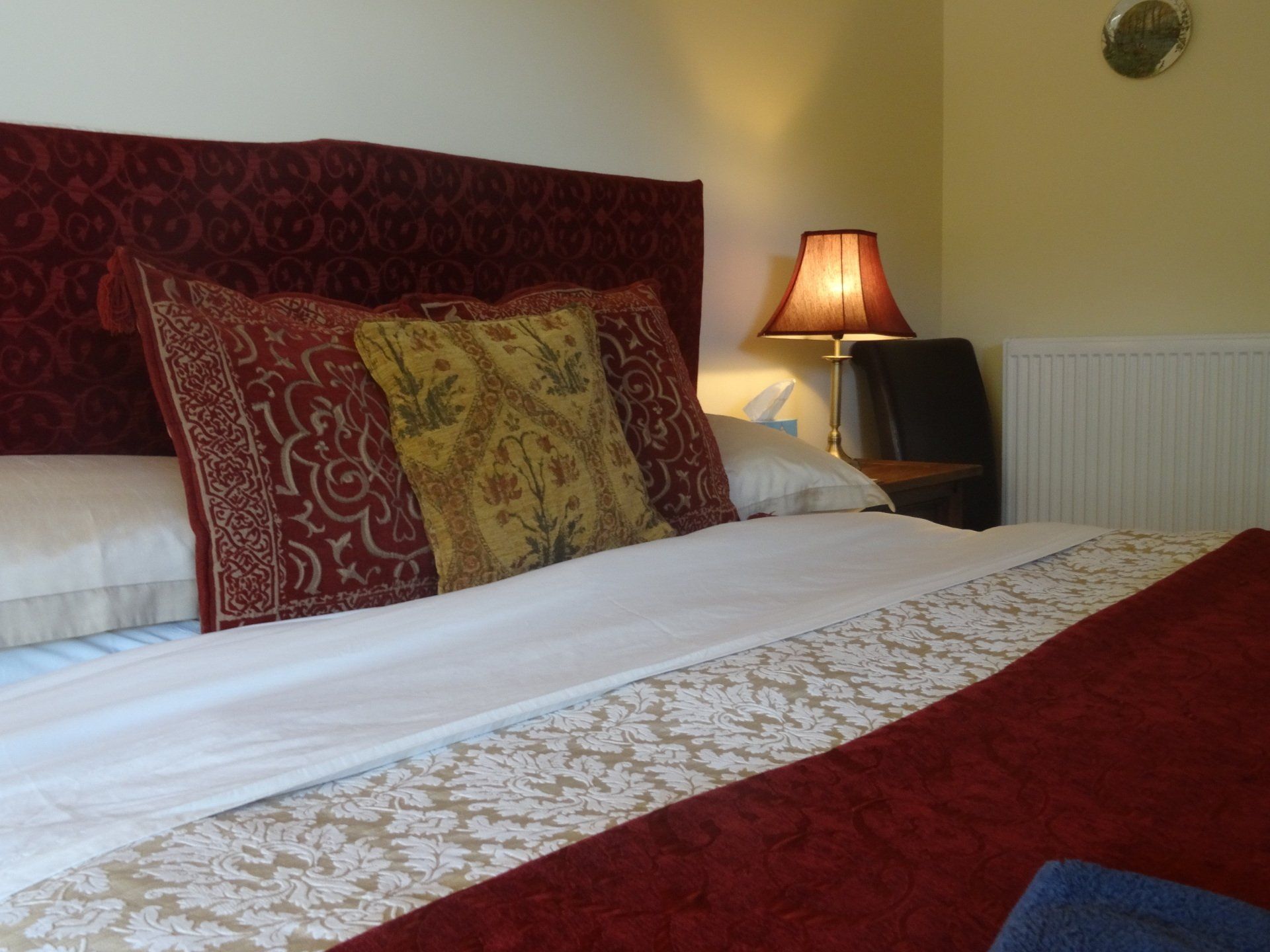 Cozy bedroom with red headboard, patterned pillows, white bedding, a lampshade, and a wall clock.