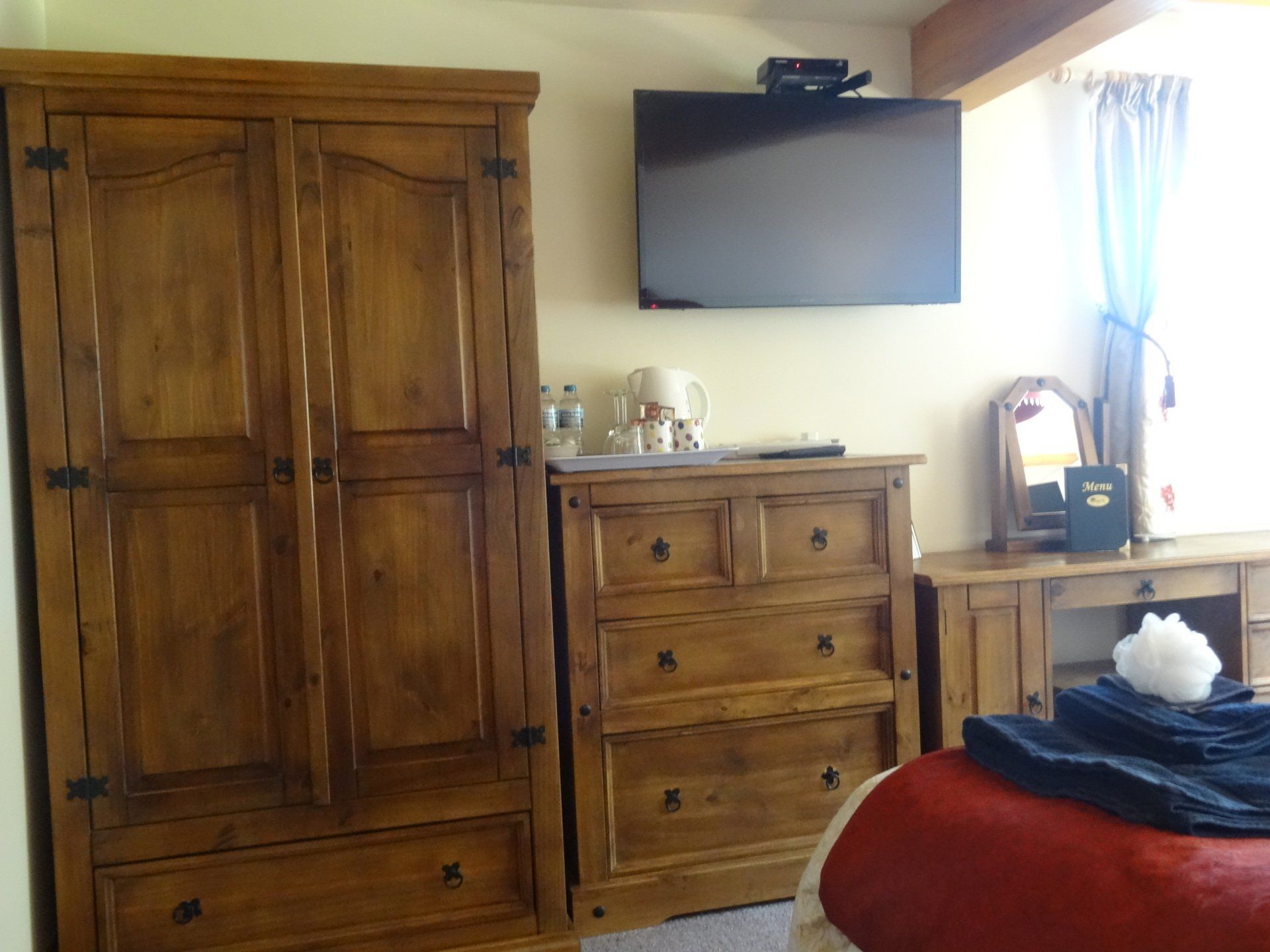 Wooden bedroom furniture including a wardrobe, chest of drawers, and vanity. A TV is mounted on the wall.