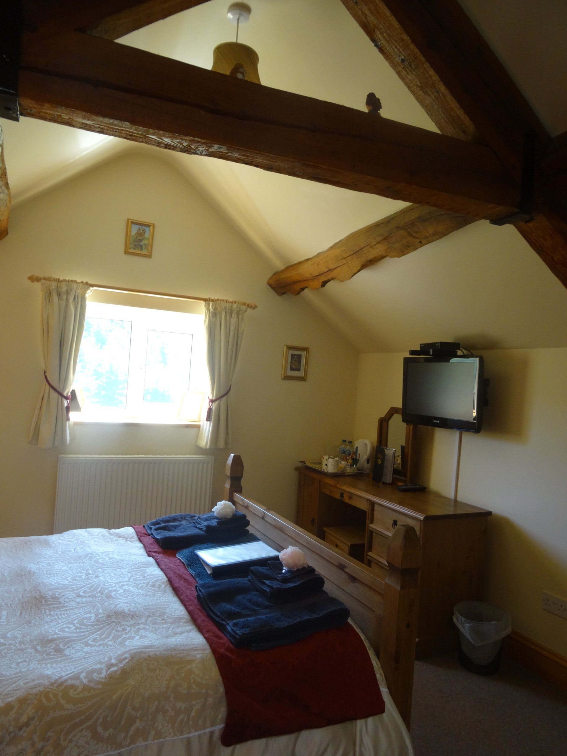 Bedroom with wooden beams, window, bed with red throw, dresser, and wall-mounted TV.