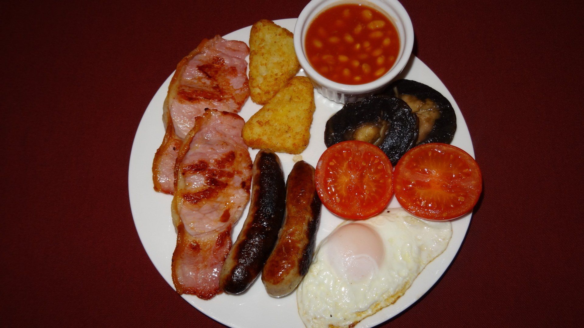 Full English breakfast: bacon, sausage, fried egg, beans, tomatoes, mushrooms, hash browns on a white plate.