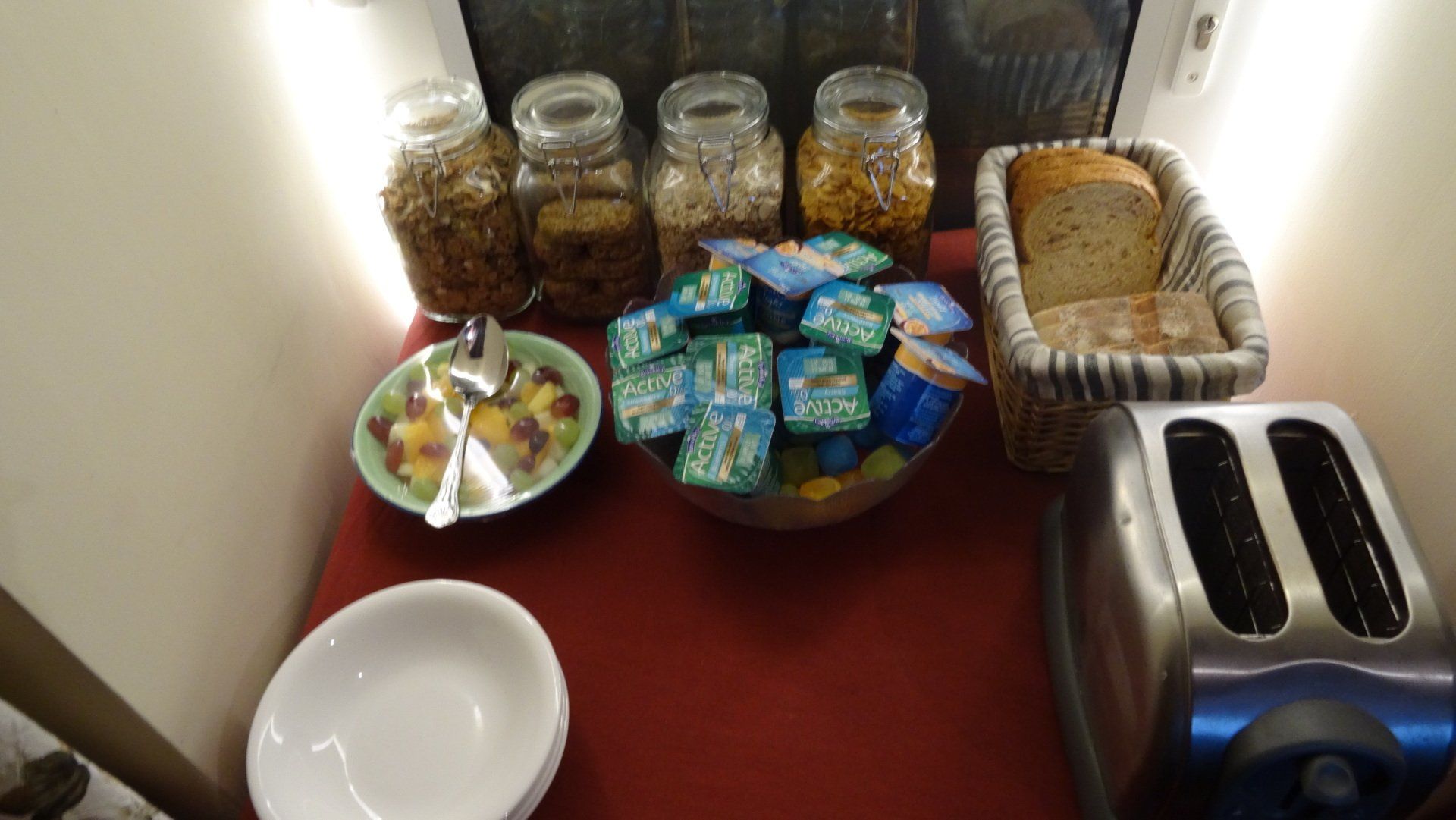 Breakfast buffet with cereals in jars, bread, yogurt cups, and a toaster.