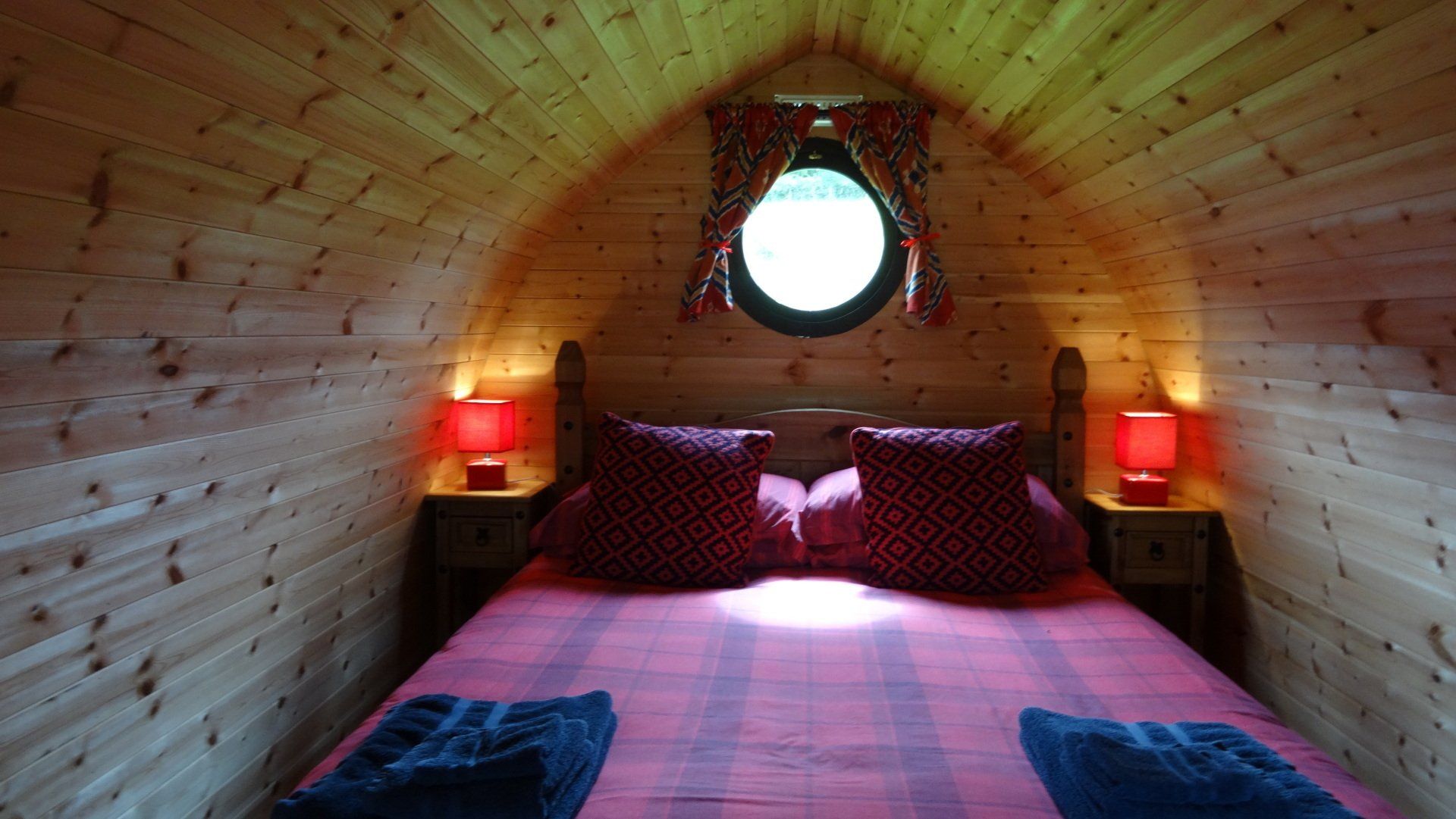 Cozy wooden pod interior with a bed, red lamps, round window with curtains.