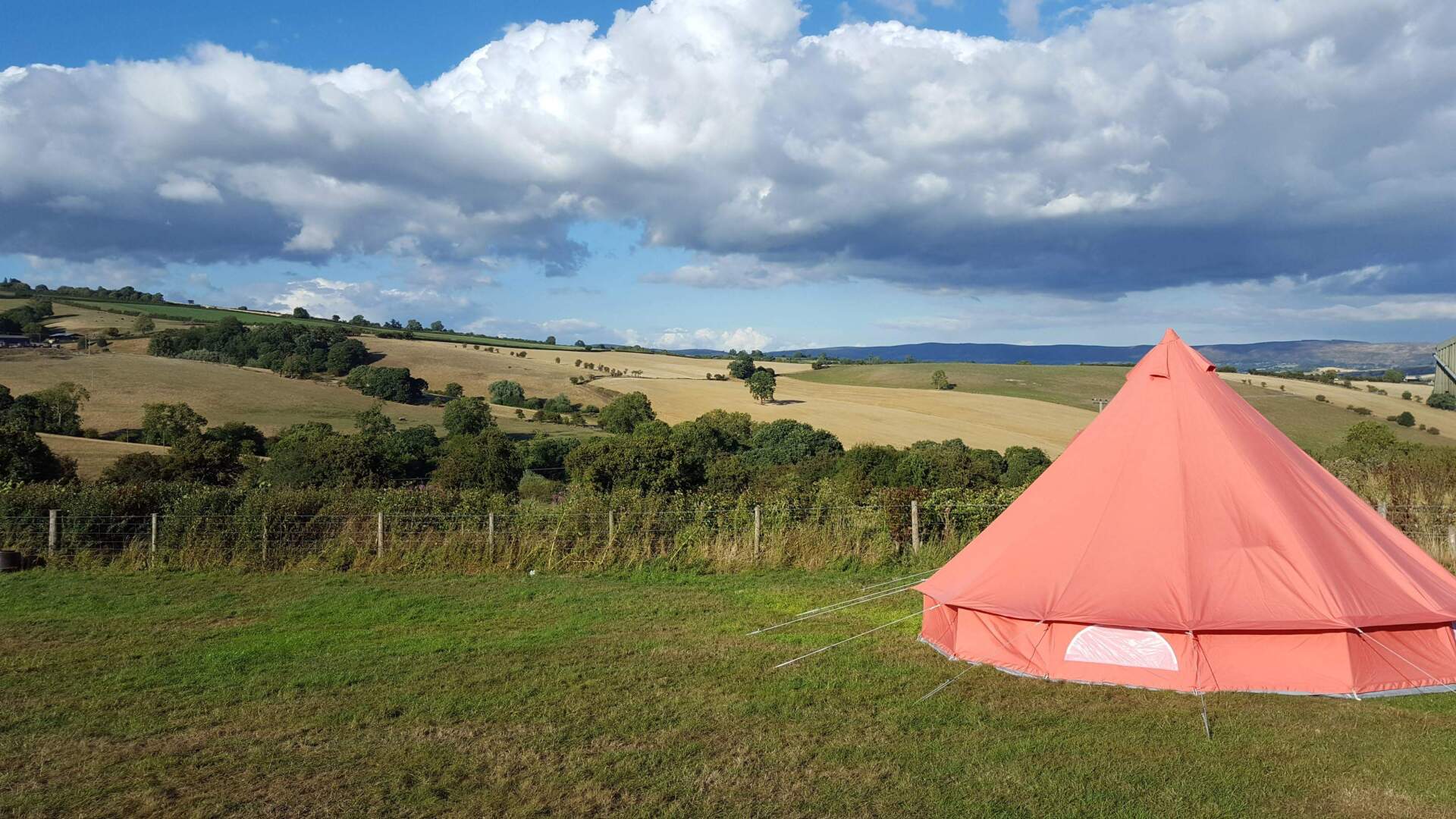 Red tent pitched in a grassy field with rolling hills and a cloudy sky.