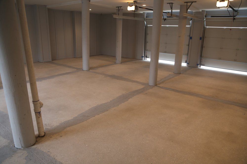 A spacious, unfinished basement with light-colored concrete floors, support pillars, and visible garage door.