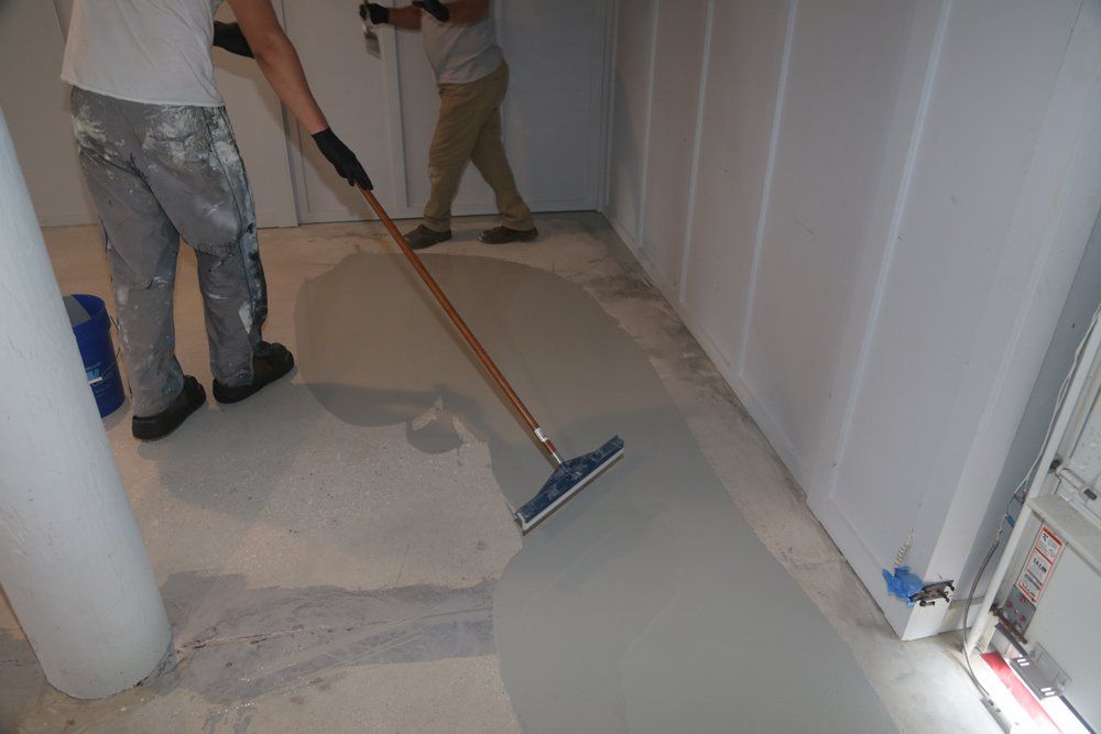 Two workers applying gray sealant to a concrete floor in a white-walled room using squeegees.
